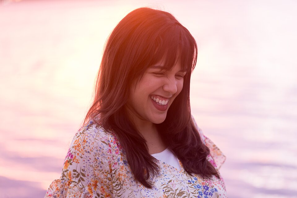 A woman laughing with her eyes closed, backlit by a pink sunset over the water