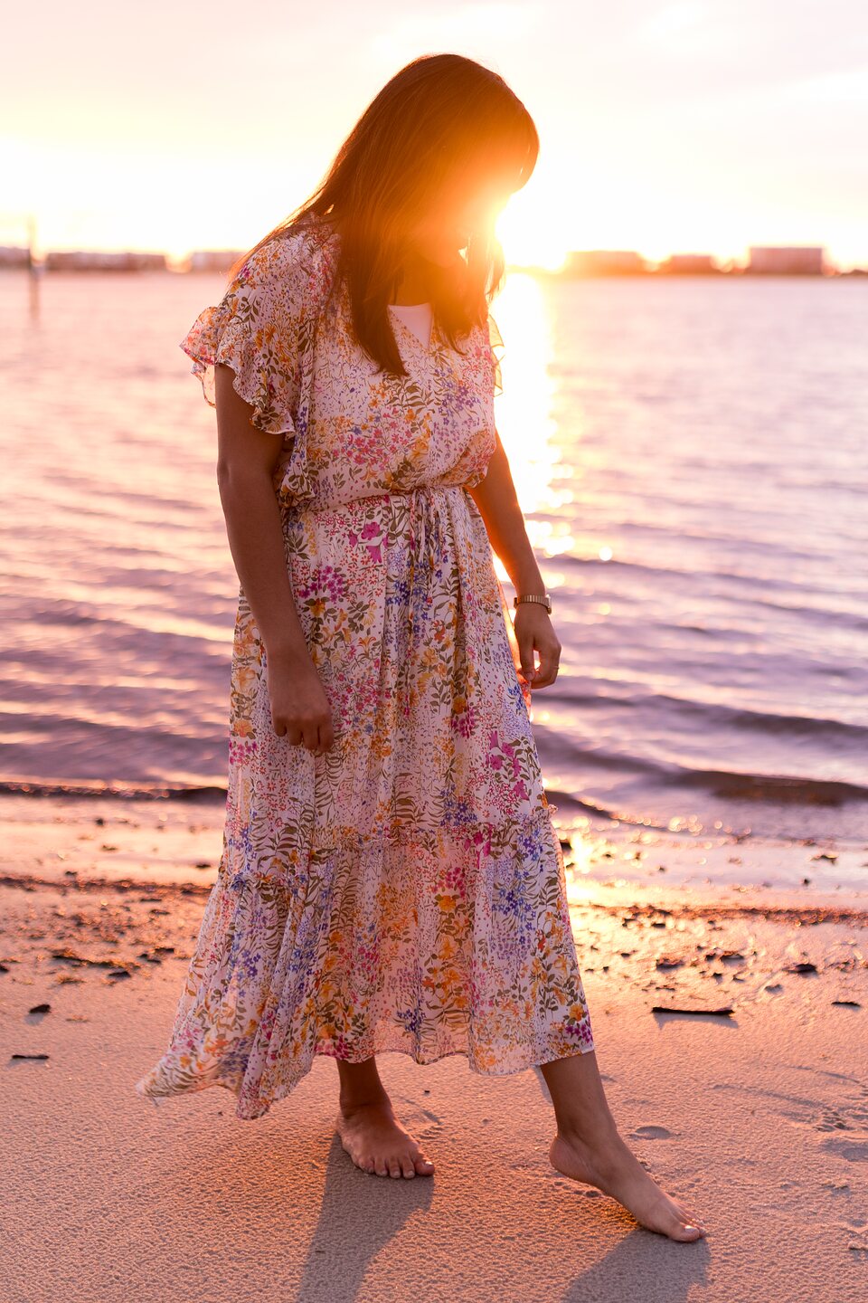 A woman in a floral dress walking barefoot on the sand as the sun sets over the water