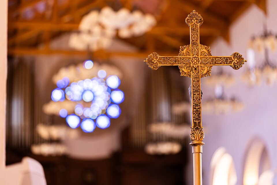 A gilded processional cross in front of the church organ, chandeliers out of focus behind