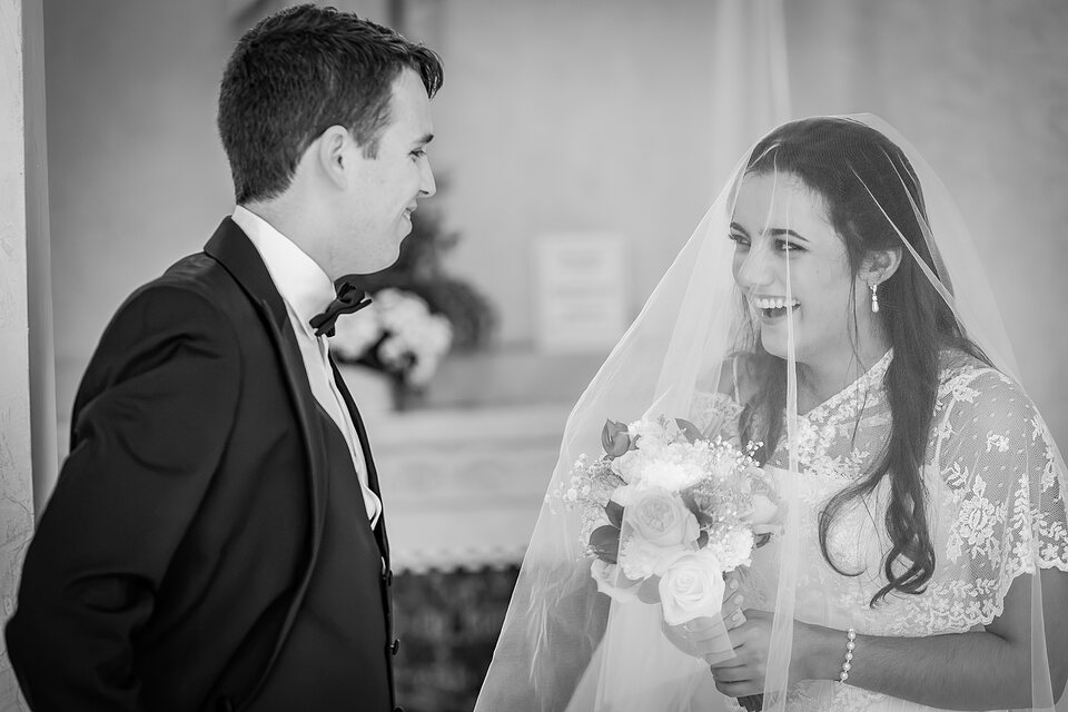 A groom and his veiled bride laughing as they face each other at the altar
