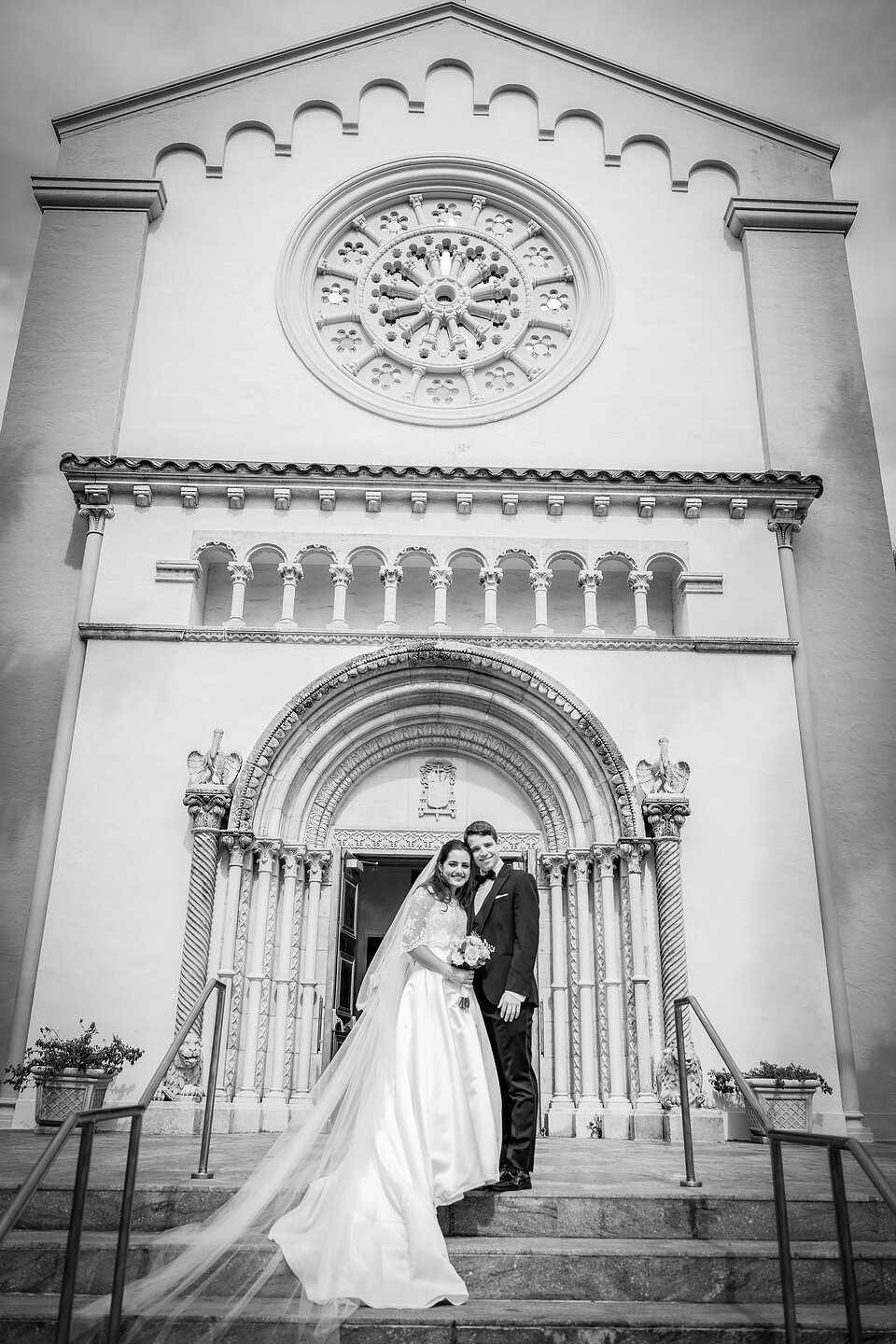 A bride and groom on the steps of a Romanesque church, her veil trailing down the stairs