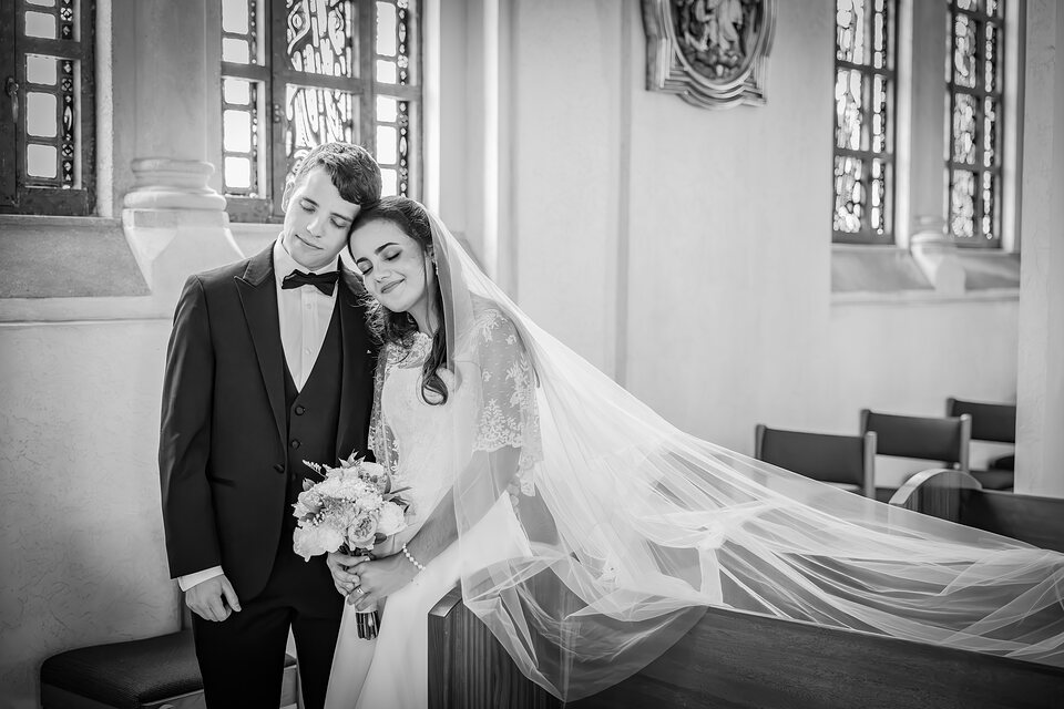 A bride resting her head on the groom's shoulder, her veil spread across the pew beside them